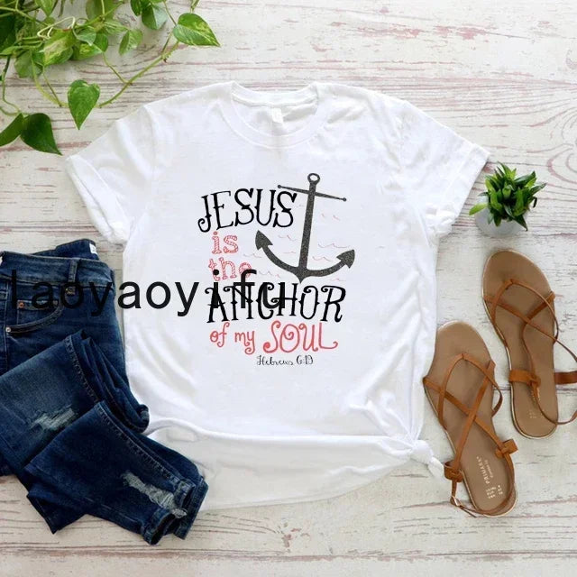 White t-shirt with 'Jesus is the anchor of my soul' design on a wooden surface with jeans and sandals.